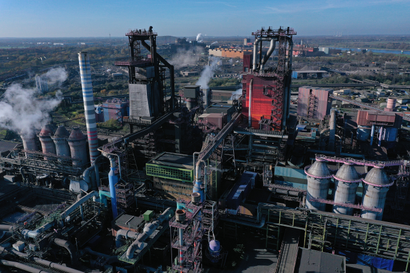 An aerial view of a German steelworks on a sunny day, with two blast furnaces and multiple towers with smoke coming from them. In the background is a river and a blue sky.