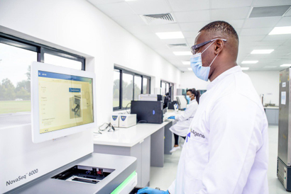 A picture of a man in a lab, wearing a white coat and a nose mask, facing a screen.