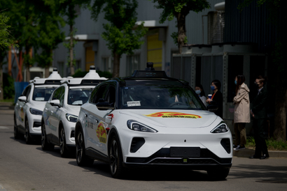 Three white autonomous vehicles (they appear to be an SUV and minivan crossover) are parked in a row on a street. A line of people stand on the side of the road facing the cars. There are trees and what appears to be a warehouse in the background.