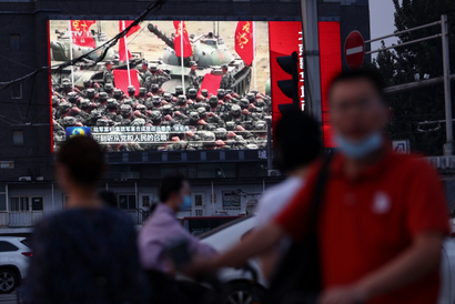 A massive screen on the side of a building displays an image of the People's Liberation Army. In the foreground are people moving, blurry. One main faces the camera wearing a red shirt and a blue face mask pulled down under his chin.
