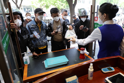 A staff member passes takeaway food to a delivery worker while others wait for their turn. Four people are waiting in the foreground to collect, all wearing white masks. Two are wearing motorcycle helmets. A man in a jean jacket and black baseball cap holds up his phone to show the restaurant staff order details.