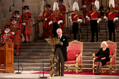 King Charles III stands in front of a golden podium delivering a speech wearing a black coat, black tie, and grey slacks. Behind him is Queen Consort Camilla on one of two red and golden chair high-backed chairs on a red carpet. There is a staircase further behind lined with men wearing an array of red and gold feathery regalia.