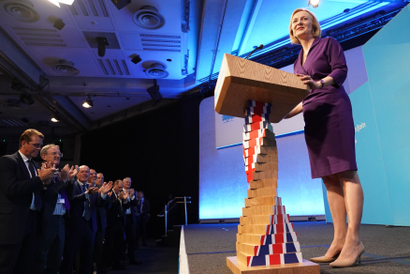 Liz Truss stands smiling on stage at a wooden podium whose column is a stylized, twisted UK flag. She is wearing a purple wrap dress and nude pumps. A row of men in suits stands in the front row before the stage clapping.