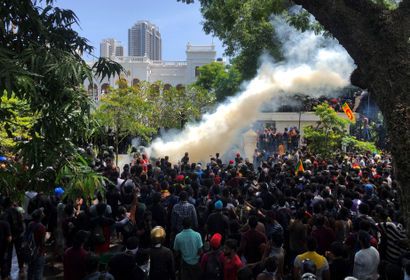 A crowd of people outside a building surrounded by trees. They're all facing a big plume of smoke.