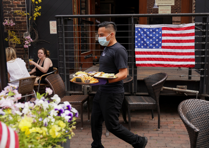 A waiter wearing a mask brings food to customers seated outside in front of an American flag
