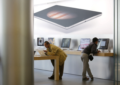 Two men lean over counters containing Apple computers, with a graphic of an iPad with a planet on it seeming to hover overtop of them but it's probably just on the wall.