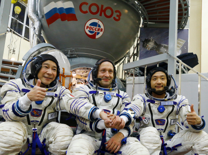 Three astronauts sit next to each other in full white and blue-accented gear space, two on each side giving a thumbs up, their hands clasped together in the center. All are smiling. In the background is some kind of gray tank with a Russian flag and lettering in what looks like a space-related facility.
