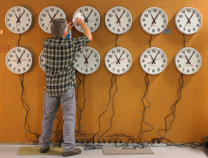 A man cleans the face of a clock hanging among 12 clocks on a wall.
