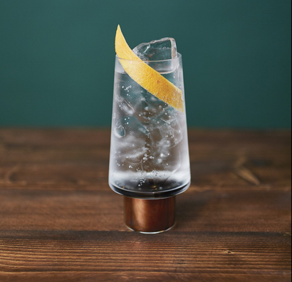 A glass of what appears to be gin and tonic on a dark wooden table on a raised coaster. There is a slice of lemon in the glass.