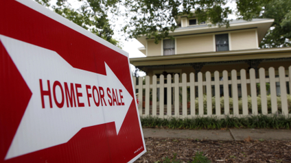 a red sign with a white arrow reads FOR SALE and sits in the yard of a yellow house with white fence
