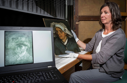A woman sits on the right side of the frame holding up the original painting, &quot;Head of a Peasant Woman,&quot; in gloved hands. In the foreground is a laptop opened with an image of the Van Gogh self-portrait x-ray conveyed in shades of blue-gray and white.