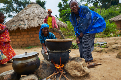 A woman laughing while she bends over a cookpot over an open fire. There are brick huts in the background with thatched roofs and three other people smiling in the picture.