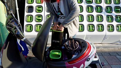 A person holds a battery over an electric scooter. There is a wall of green-handled batteries behind them.