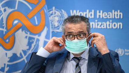 World Health Organization Director-General Tedros Adhanom Ghebreyesus adjusts his face mask while seated in front of a banner with the WHO logo.