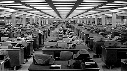 A black and white gif from an old movie of a man working alone in a large open-plan office.