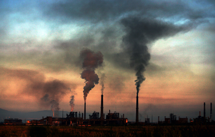 A photo that almost looks like a watercolor, of the silhouette of smoking chimneys at a steel plant against a setting (or rising sun). Glowing fires can be seen also against the dark buildings and equipment.