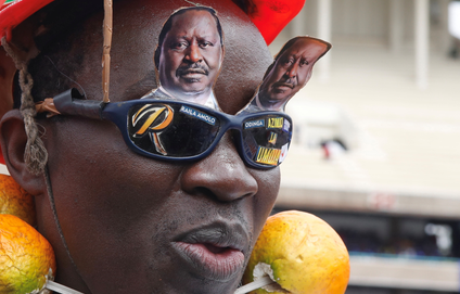 A portrait of a man wearing black-lens sunglasses with a navy frame. On top of the glasses are two paper cutouts of Raila Odinga. On the frames are more paper cutouts including a large, orangey-yellow letter "R" and the words "Raile Amolo Odinga." Around the man's neck appears to be some kind of giant faux-fruit (mango?) necklace. He is also wearing a red helmet.