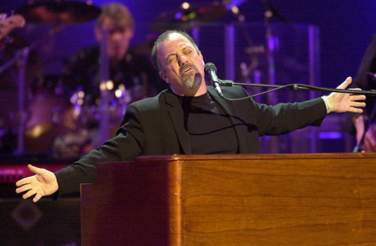 Singer Billy Joel performs at a piano with his arms outstretched. 