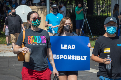 Three workers wearing cloth masks walking out of their job at Blizzard. One is holding a sign that reads "Lead Responsibly"