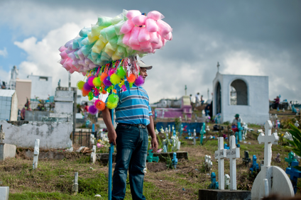 Un hombre sostiene un palo con bolsas de algodón de azúcar y juguetes misteriosos en equilibrio sobre él.  Él está de pie en un cementerio. 