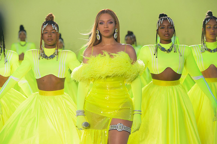 Beyonce and several dancers wearing neon yellow-green outfits that are very fun. Beyonce is holding a microphone and the wind is in her hair.