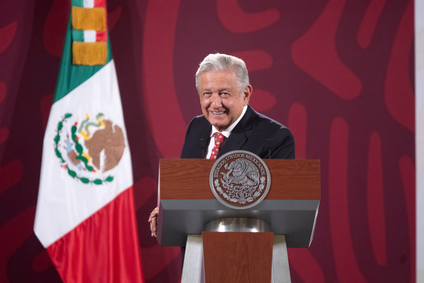 Mexican President Andres Manuel Lopez Obrador at a podium with the Mexican seal on it, in front of the Mexican flag. He is leaning casually on the podium and smiling.