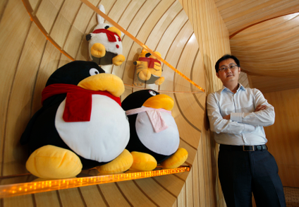 In the foreground are two giant, stylised penguin plushies with red and pink scarves, respectively, on an orange-tinged glass shelf, above it is another shelf with two more plushies that are slightly smaller, also with scarves. One looks like a rabbit, the other is a tiger. Tencent&#039;s CEO is in the back, leaning against the wall and looking slightly down at the cameraman with his arms crossed. The space is covered floor to ceiling in light-colored wood panels, some of it curved concave and convex.