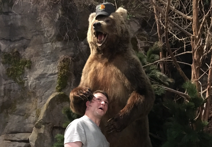 A man with an ironically scared expression on his face has put his head between the claws of a taxidermied bear wearing a baseball cap.