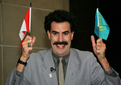 Actor Sacha Baron Cohen holds two mini flags and is aggressively smiling. 