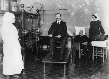 A black and white photo of nurses with large dresses and nun-like habits around their heads, watching a man sitting on a chair, on a table, with a large mustache, hold onto some kind of rod attached to a large machine. Everyone looks surprisingly relaxed.