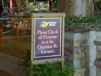 A sign saying Please check all firearms in at the Cherokee St. Entrance in front of a Bass Pro Shop lit up with holiday decorations.