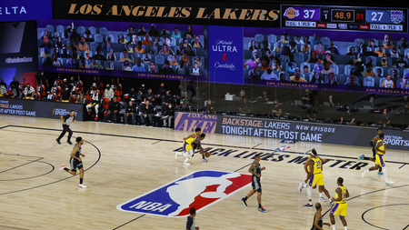 Two teams play in a nearly empty arena in front of a large video screen displaying the heads of virtual fans.