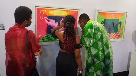 An artist at Art X Lagos 2021 explains her work to two guests on her left and right. The person on the right is looking closely at the painting on the wall.