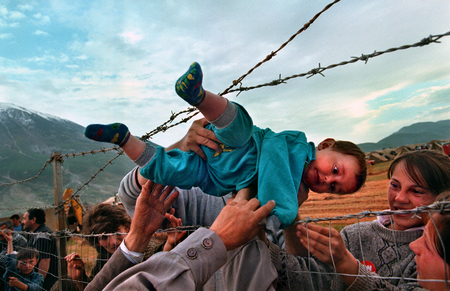 Agim Shala, 2 years old, is passed thru the barbed wire fence at a refugee camp as members of the Shala family are reunited after fleeing Kosovo, in 1989. The relatives who just arrived from Prizren had to stay outside the camp until shelter was available.