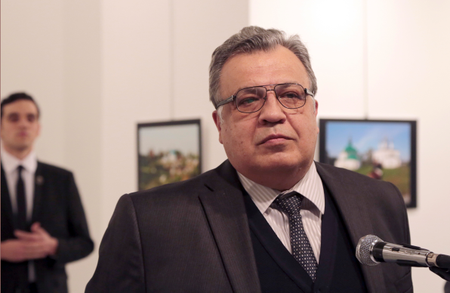 Andrei Karlov, the Russian Ambassador to Turkey, speaks at a photo exhibition in Ankara on Monday, Dec. 19, 2016, moments before a gunman opened fire on him. Karlov was rushed to a hospital after the attack and later died from his gunshot wounds. The gunman is seen at rear on the left. (AP Photo/Burhan Ozbilici)