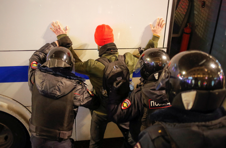A man in a jacket and orange skull cap has is hands against a white vehicle while police in black helmets put their hands on him to search and arrest him