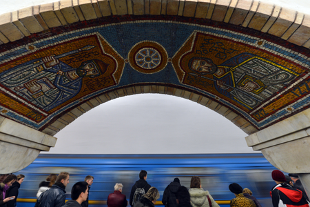 Mosaic tiles in red blue and y ellow form a design over an archway in a subway station. Below the arch are passengers waiting for a blue subway train.