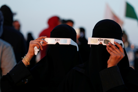 Saudi women holding protective glasses over black burkas that cover them completely watch the eclipse