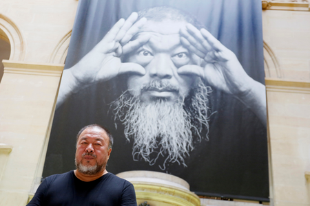 Ai Weiwei poses under the poster of an exhibition titled &quot;Ai Weiwei: By the way, it&#039;s always the others&quot; in Switzerland.