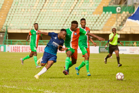 A soccer player for Sporting Lagos in a blue jersey competes for the ball with a Go Round FC player in a green and orange Jersey.