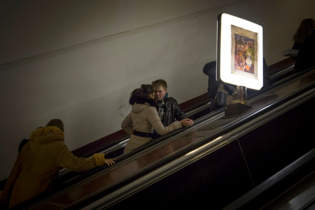two people speak while riding an up escalator in an underground subway station. A flourescent light with a picture decorating it lights the escalator.