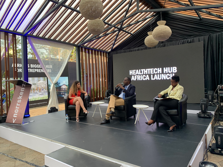 A man and two women sit on chairs. Behind them is a screen with the words &quot;HEALTHTECH HUB AFRICA LAUNCH&quot; in white.