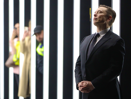 An image of Elon Musk in a black suit and dark tie, standing with his hands clasped in front and head raised, eyes closed. In the background are consecutive vertical white and black bars, three blurry figures stand in between them. 