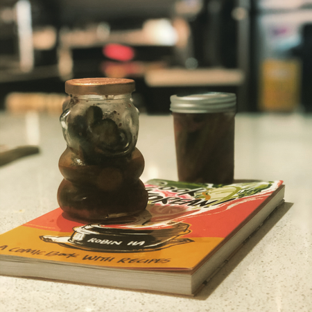 A jar of pickles in a bear-shaped jar and a jar of pickled okra on a counter top. They are on top of a cookbook by Robin Ha,