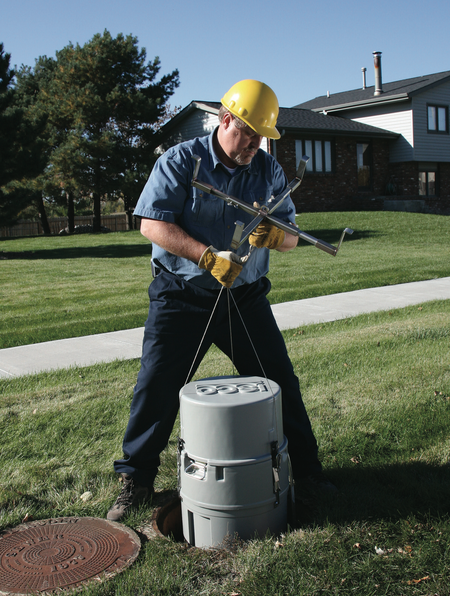 A man working outside wearing a hardhat. He&#039;s carrying a cylindrical autosampler that is about 2 feet long and a foot wide.