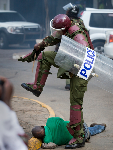 A Kenyan riot policeman repeatedly kicks a protester as he lies in the street.