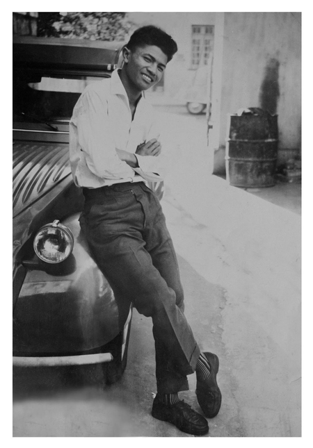 A black and white image of a young Malagasy man leaning against a car
