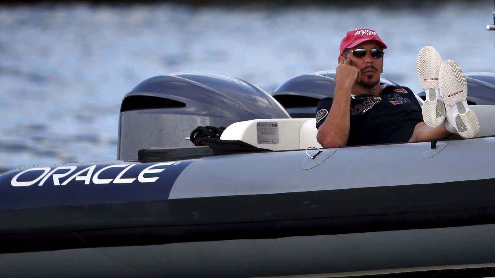 Larry Ellison reclines with his feet up in a boat, wearing sunglasses and a red cap.