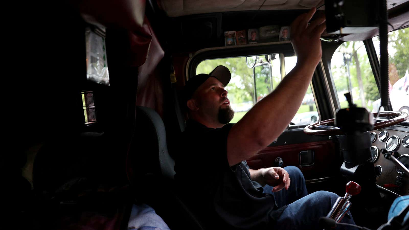 A man with a beard and a baseball cap sits in the cab of his truck.