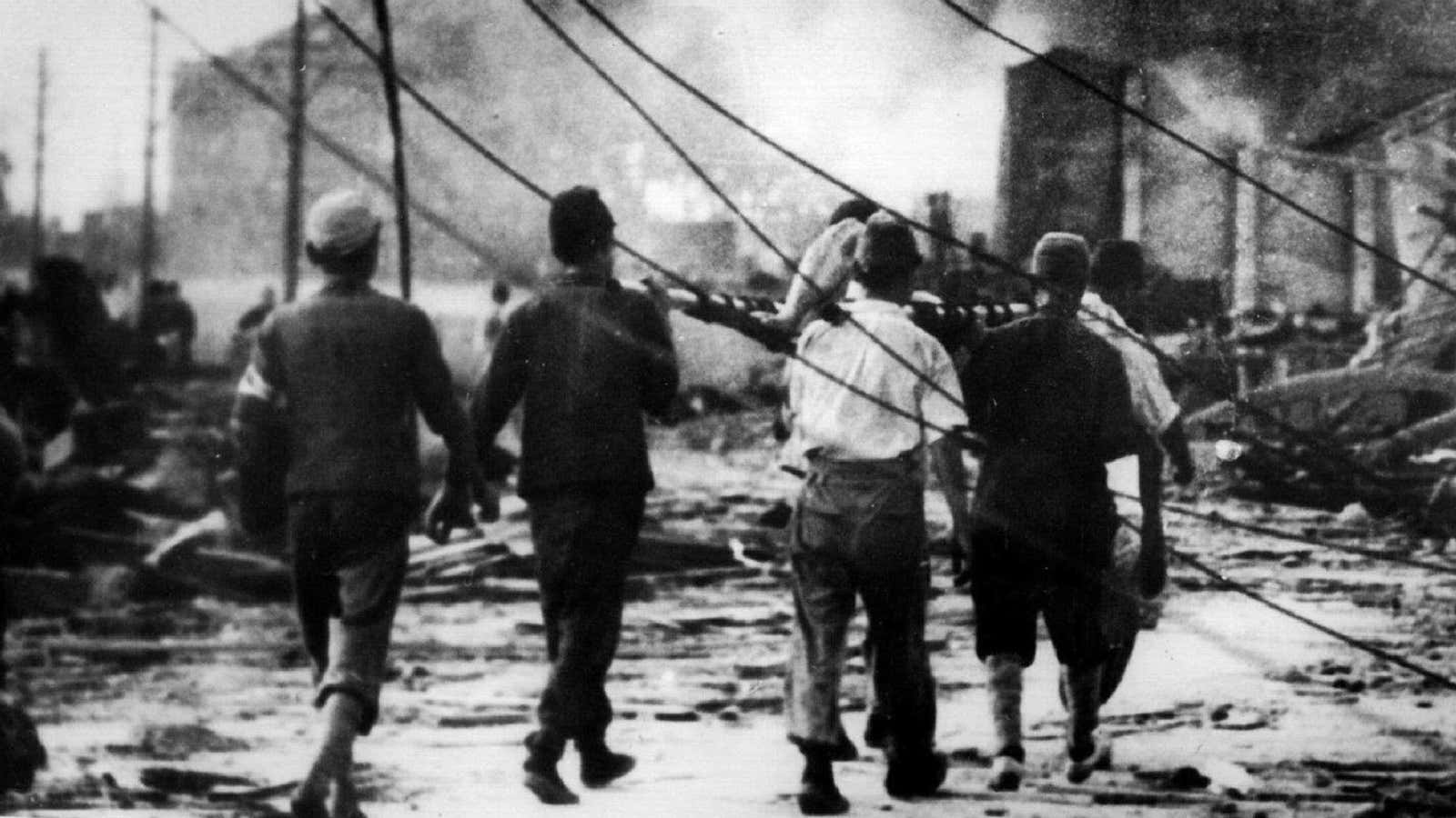 Japanese air raid workers carry a Hiroshima victim away from smoking ruins.